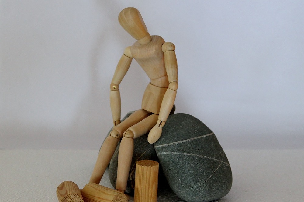 Figurine articulée en bois assise sur un caillou, dans une posture de grande fatigue