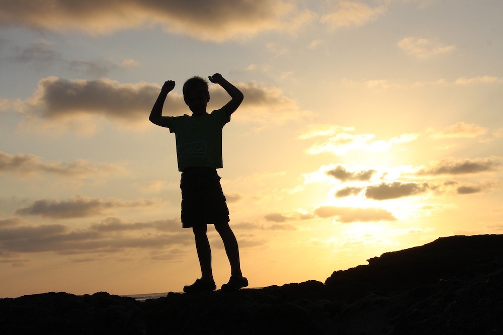Un enfant debout sur une crête, les bras levés en signe de victoire, en contre-jour devant un coucher de soleil